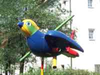 Sparrow sculpture with blue body and beak, red tail, yellow and green head, and primary-color pipes piercing it
